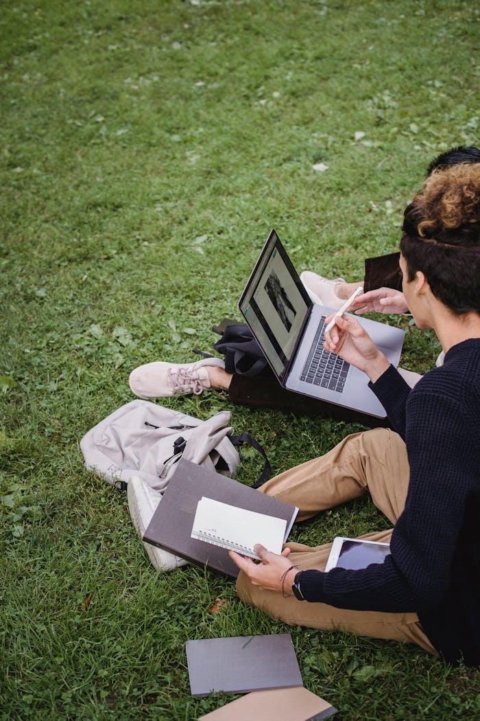 Two students study together on a laptop outdoors, surrounded by books.